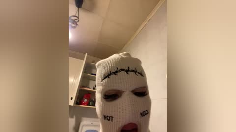 Video of a woman wearing a white knitted beanie with \"NOT\" and \"KILL\" written on it, inside a small, dimly-lit room with beige walls, a wooden shelf holding red items, and a ceiling light.