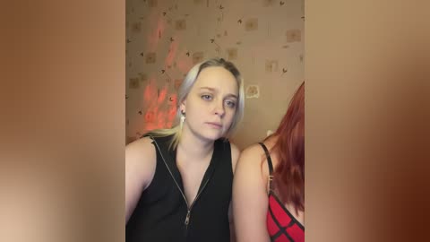 Video of a young woman with long blonde hair and fair skin, wearing a black zip-up top, standing beside a redhead in a red and black plaid shirt. Background features a beige wallpaper with floral patterns and red light effects.