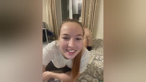 Video of a smiling, fair-skinned woman with long, straight brown hair, lying on a bed with floral-patterned sheets, wearing a white t-shirt, and a partially visible person in the background.