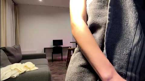 Video of a person in a gray, textured bathrobe standing in a modern, minimalist living room with beige curtains, wooden floor, and a TV on a table.