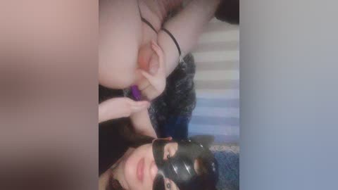 A blurry, low-quality video shows a close-up of a woman in a black mask, with her breasts exposed, being held by another woman's hands, both indoors.