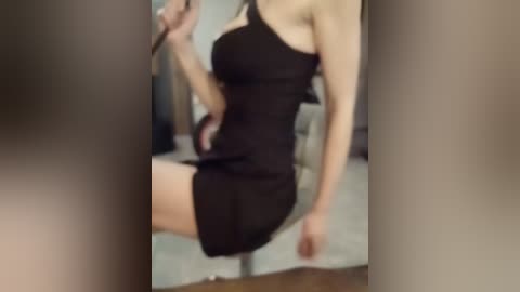 A video of a slender, fair-skinned woman in a black sleeveless dress, captured in a mirror, with blurred background.