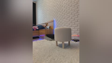 Video of a modern bedroom with a white brick wall, wooden bed with pink bedding, beige armchair, and a fluffy white carpet.