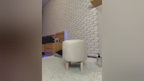 Video of a modern, minimalist bedroom with a white brick wall, light wooden furniture, and a beige ribbed ottoman on a white shag carpet.