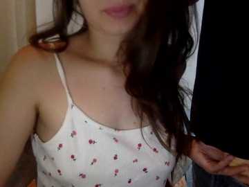 Video of a young woman with fair skin and long brown hair, wearing a white spaghetti-strap tank top adorned with small red flowers. She is indoors, partially obscured by a dark curtain in the background.