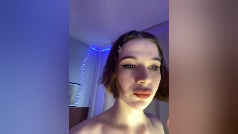 Video of a fair-skinned woman with short brown hair, wearing black eyeliner and red lipstick, standing in a dimly lit room with blue LED strip lights.
