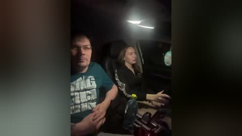 Video of a dark, dimly-lit car interior featuring two people in the backseat. The man on the left wears a blue \"BEASTIE BOYS\" t-shirt, and the woman beside him has long hair and a black jacket.