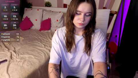 Video of a young woman with long brown hair, wearing a white t-shirt, sitting on a bed with beige sheets and pink pillows. A video game screen overlays the image.