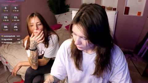 Video of two tattooed women in casual white T-shirts, one with long brown hair, the other with long blonde hair, sitting on a bed in a cozy bedroom.