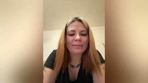 A video of a fair-skinned woman with long, straight, light brown hair, wearing a black V-neck shirt and a black necklace with a floral design. She has a slight smile and is looking down. Background features beige walls.