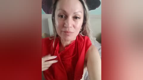 A video of a fair-skinned woman with long, straight blonde hair, wearing a red sleeveless top, and a black hat, posing indoors with blurred faces on either side.