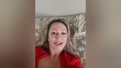 Video of a Caucasian woman with shoulder-length blonde hair, fair skin, and a slight smile, wearing a red top, reclining on a pillow with newspaper print.