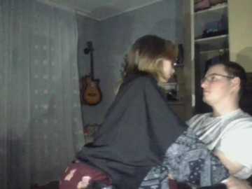 A video depicts a woman with blonde hair wearing a black shirt and maroon pants, sitting on a bed in a dimly lit room with a guitar on the wall, and a man with glasses wearing a gray shirt and patterned pants, sitting nearby.