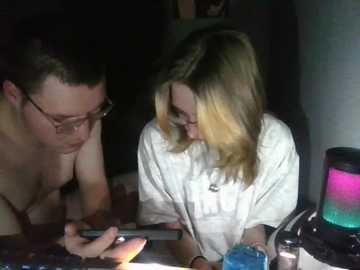 A dimly lit video of two people: a man with glasses, shirtless, and a woman with blonde hair, both focused on a smartphone. The background is dark with a glowing lava lamp.