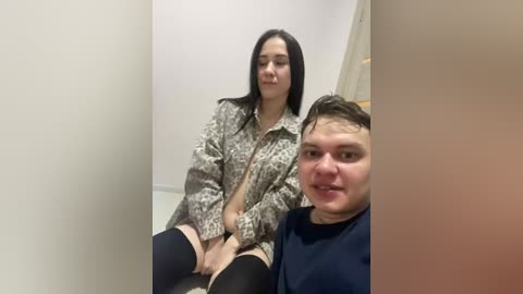 Video of a woman with long black hair, wearing a patterned jacket and black stockings, sitting next to a man in a blue shirt, both in a dimly lit, beige-walled room.