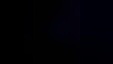 A dimly lit, grainy black and white video features a dark silhouette of a person standing on the right side, partially obscured by shadows. The background is predominantly black, with no discernible objects or textures.