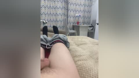 Video of a man's bare legs with socks, lying on a beige blanket in a room with zigzag-patterned curtains and a small table.