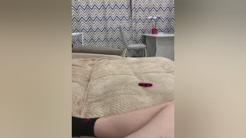 Video of a beige, textured quilt with a red object on it in a room with blue and white zigzag-patterned wallpaper and a white chair.