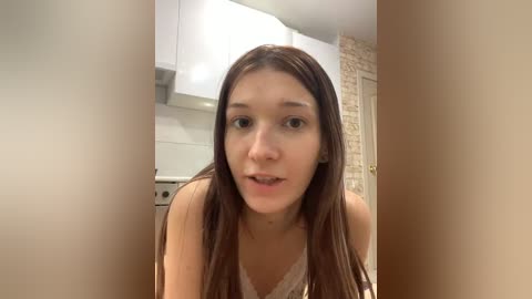 A video of a young woman with long brown hair, wearing a white lace bralette, standing in a modern kitchen with white cabinets and tiled backsplash.