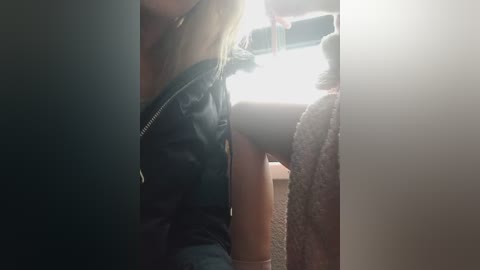 Video of a blonde woman in a black leather jacket, leaning against a beige wall, with a white towel hanging beside her.