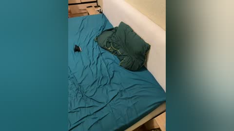 A video showing a simple, unmade bed with teal sheets and a green pillow, against a beige wall in a small, sparsely furnished room.