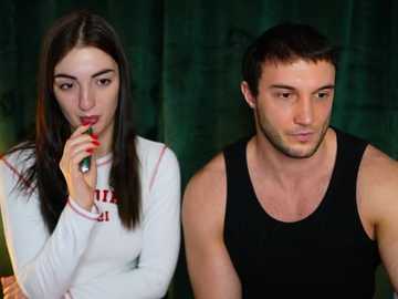 Video of a young woman with long dark hair, wearing a white long-sleeve shirt, and a young man with short dark hair, wearing a black tank top, both sitting against a dark green backdrop.