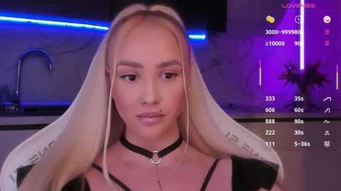 Video of a fair-skinned woman with long blonde hair, wearing a black choker and silver nose ring, in a modern kitchen with purple lighting.