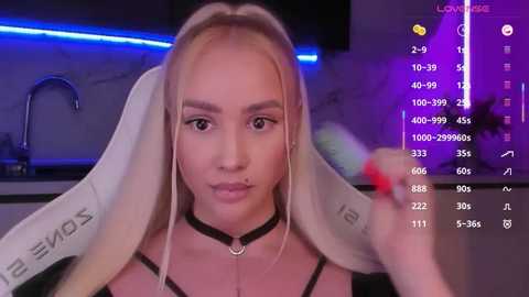 Video of a young, fair-skinned, blonde woman with long hair, wearing a black choker and black top, in a modern kitchen with purple lighting, holding a paintbrush.