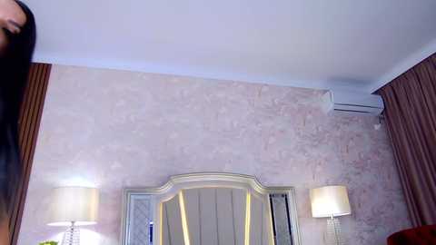Video of a plush, modern hotel room with a light beige marble wall, white lampshades, and an air conditioner on the wall.