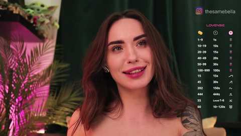 Video of a smiling, attractive woman with long, dark hair and a tattooed arm, wearing pink lipstick, in a tropical-themed room with a green curtain and a plant in the background.