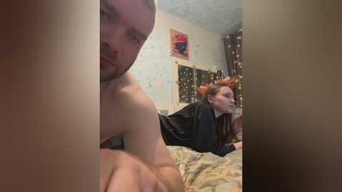 Video of a young couple in bed; man shirtless, woman with long hair, black robe, and bed sheets; room with patterned wallpaper, fairy lights, and a red poster.