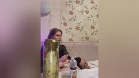 Video of a woman breastfeeding in a hospital room with a floral wallpaper background. She's holding a baby, with a green ceramic pitcher and a bottle nearby.