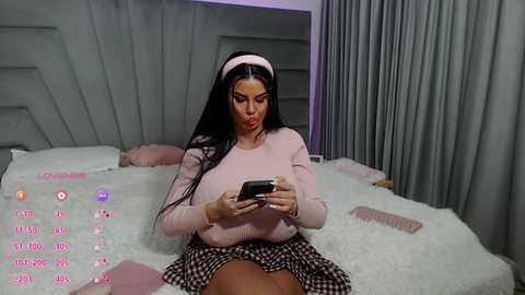 A video shows a Latina woman with long black hair, wearing a pink headband and checkered skirt, sitting on a fluffy white bed, playing with a smartphone.