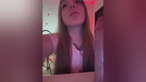 Video of a fair-skinned woman with long, straight brown hair, wearing a pink top and a feather necklace, taking a selfie in a pink-themed bedroom with red butterfly decals and fairy lights.