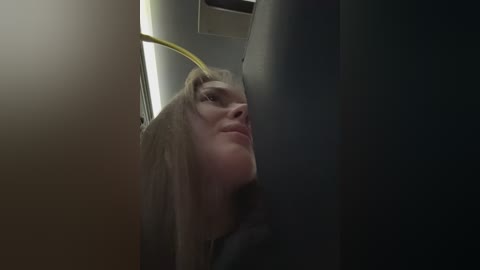 Video of a young woman with long, blonde hair, wearing a gray jacket, standing in a dimly lit, narrow hallway with a yellow light fixture overhead.