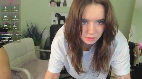 A video of a young woman with long brown hair, wearing a white t-shirt, in a modern living room with green walls, black leather furniture, and a potted plant.