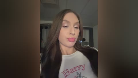 Video of a young Caucasian woman with long brown hair, light skin, and pink lips, wearing a white \"Berry\" t-shirt, indoors with dim lighting.