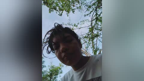 Video of a young woman with short, curly dark hair, wearing a white t-shirt, smiling against a backdrop of green leaves and a partly cloudy sky.
