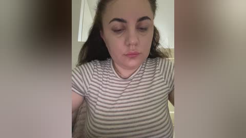 Video of a young woman with light skin, long brown hair, and closed eyes, wearing a striped t-shirt, standing indoors with a blurred background.