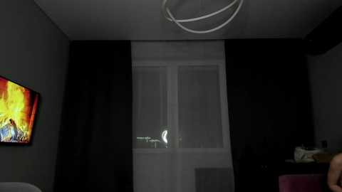 Video of a dimly lit room with a flat-screen TV displaying a colorful image on the left wall, black curtains, a white-framed window, a modern circular ceiling light, and a pink couch.