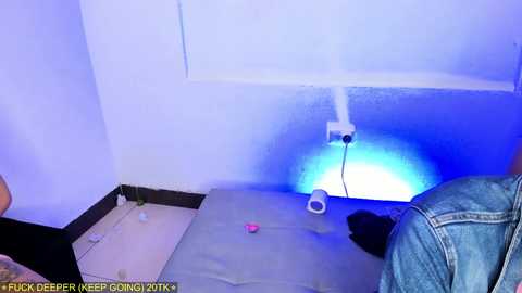 Video of a dimly-lit, cramped room with a blue glow emanating from the baseboard. A person in a denim jacket is seated on a grey mat, surrounded by scattered objects and a pink object.