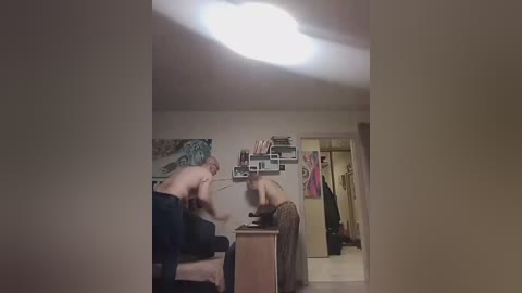 Video of a dimly-lit bedroom with two shirtless men, one seated, the other standing, a woman in a brown dress, and a cluttered wall with photos and artwork.