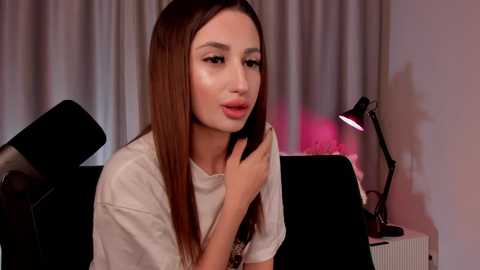 Video of a woman with long, straight brown hair, wearing a white T-shirt, sitting on a black chair, indoors with gray curtains, a desk lamp, and a pink glow.