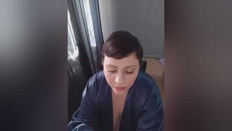 Video of a fair-skinned, short-haired woman in a dark robe, sitting on a bed, with blurred curtains and a wooden floor in the background.