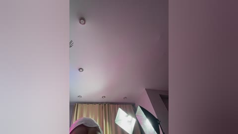 Video of a modern, minimalist room with white walls, a large window with yellow curtains, a flat-screen TV, and a person partially visible in the foreground wearing a pink and white hat.