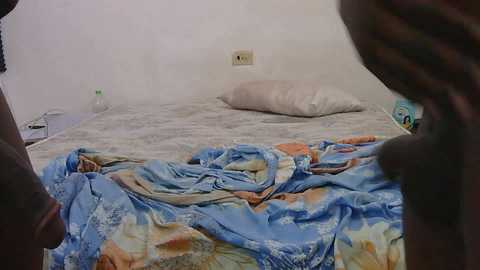 Video of a messy, unmade bed with a blue and white patterned sheet, a beige pillow, and a white wall with an electrical outlet. Two dark-skinned legs are visible at the bottom edges, suggesting a person lying on the bed.