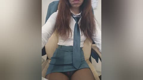 Video of a young woman with long brown hair, wearing a white blouse, blue tie, and blue pleated skirt, seated in a beige chair. Background includes a wall with a white outlet.