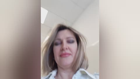 Video of a blonde woman with light skin, wearing a white blouse, smiling. Background shows a white ceiling with fluorescent lights and blurred walls.