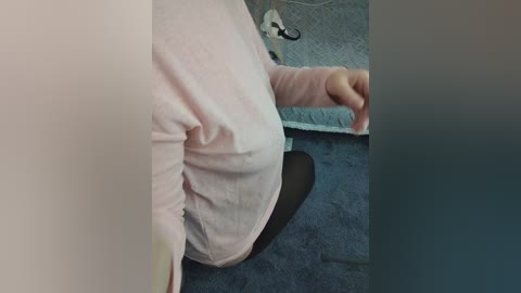 Video of a person in a light pink shirt, black tights, seated on a blue carpeted floor, with a visible light-colored blanket in the background.
