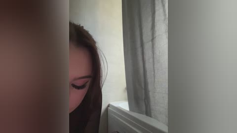 Video of a woman with long, dark hair partially obscured by a shower curtain, revealing her eye and part of her face. She appears to be in a bathroom with white walls and a grey shower curtain.